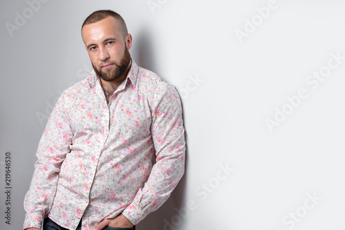 Confident looking bearded business man with short haircut, trendy dressed in white shirt with floral print looking at camera leaning on the white wall photo