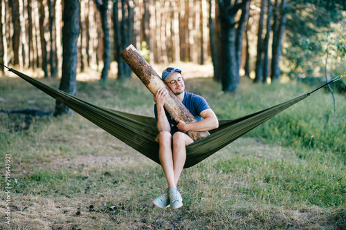 Odd weird strange unusual male person. Outlandish funny crazy foolish man sleeping in hammock with huge wooden log at nature among trees.  Wood and forest lover. With beam in bed.  Leisure lifestyle.