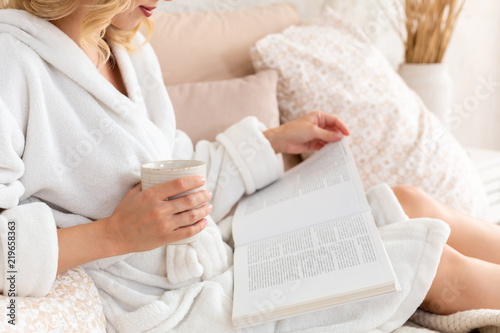 Young woman in white terry robe is drinking coffee and reading magazine or book in bedroom. photo