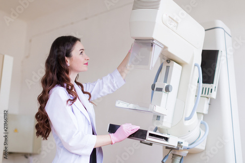 Side view of the young cheerful breast specialist in pink gloves who is preparing the apparatus of the ultrasound examination of the breast for the ultrasound examination in her office