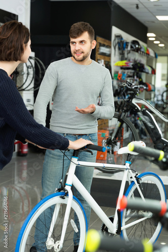 Young man seller demonstrating bicycle to woman client in store