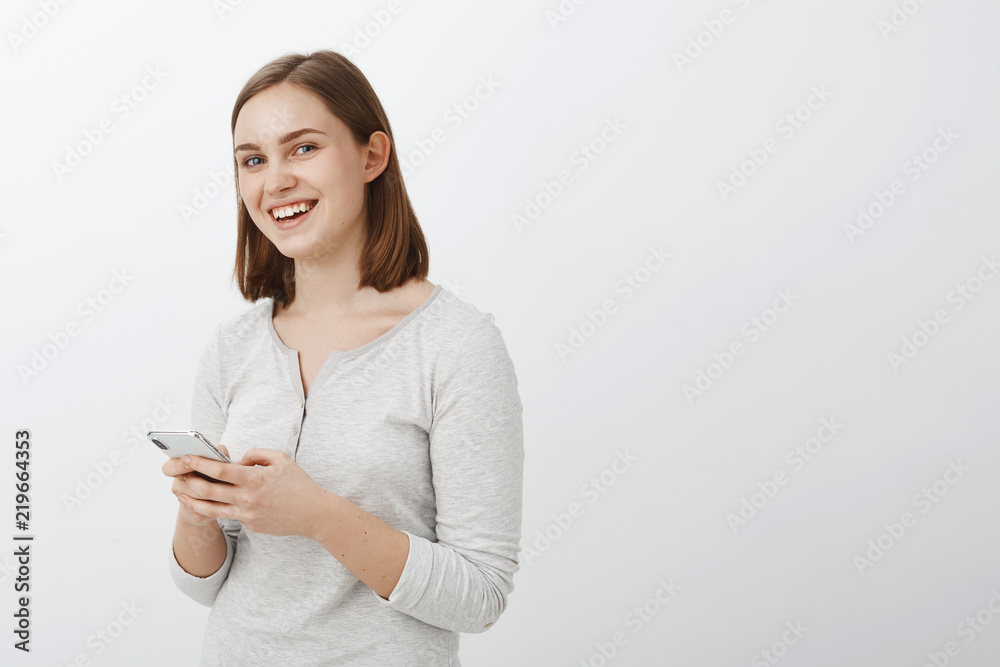 Waist-up shot of energized creative and delighted friendly-looking brunette female in casual blouse standing half-turned over gray wall holding smartphone laughing being entertained and amused Waist-up shot of energized creative and delighted friendly-looking brunette female in casual blouse standing half-turned over gray wall holding smartphone laughing being entertained and amused