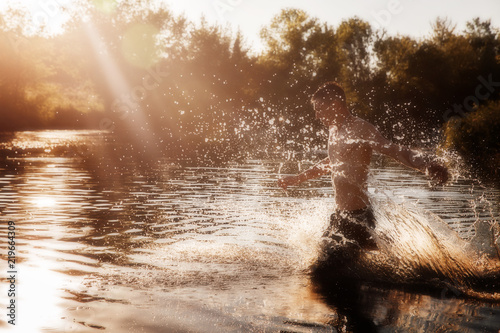 a young man with a bare torso in black shorts running through the water with splashes against the sunset