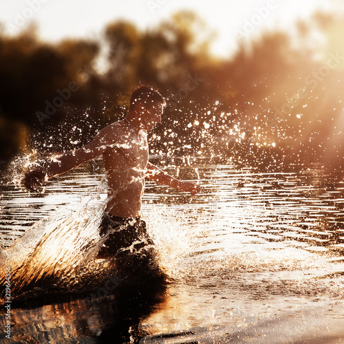 a young man with a bare torso in black shorts running through the water with splashes against the sunset