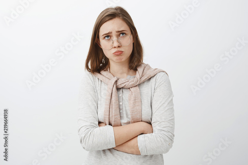 Sad gloomy offended silly girl in glasses and cute outfit folding lips, pouting frowning and looking up while complaining being offended whining over cruel and unfair life over gray background