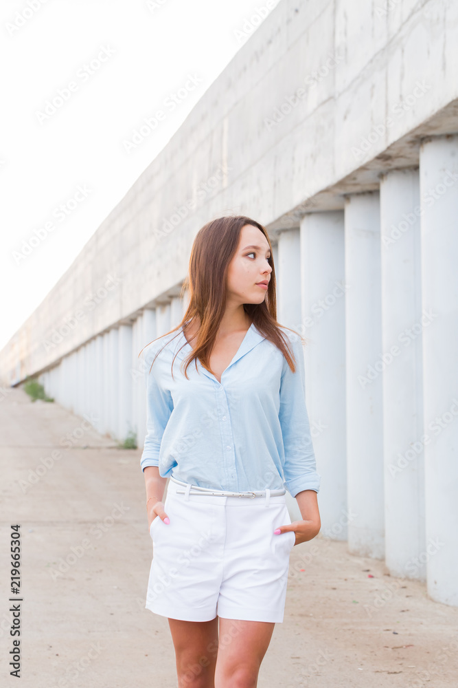 Beautiful young woman in blue shirt and white shorts walks outside in summer