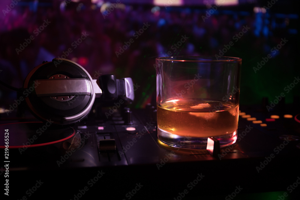 Glass with whisky with ice cube inside on dj controller at nightclub. Dj Console with club drink at music party in nightclub with disco lights. Glass with whisky with ice cube inside on dj controller at nightclub. Dj Console with club drink at music party in nightclub with disco lights.