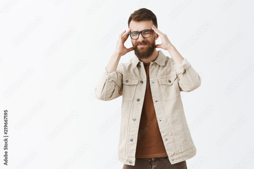 Indoor shot of intense displeased european guy with long beard in glasses and outdoor jacket having painful feeling in head holding hands on temples closing eyes and frowning from headache or migraine Indoor shot of intense displeased european guy with long beard in glasses and outdoor jacket having painful feeling in head holding hands on temples closing eyes and frowning from headache or migraine