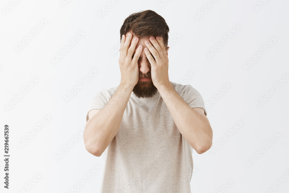 Studio shot of man suffering from painful feelings griefing bending head down covering face with palms being fed up and depressed, standing tired after working all night over gray background Studio shot of man suffering from painful feelings griefing bending head down covering face with palms being fed up and depressed, standing tired after working all night over gray background