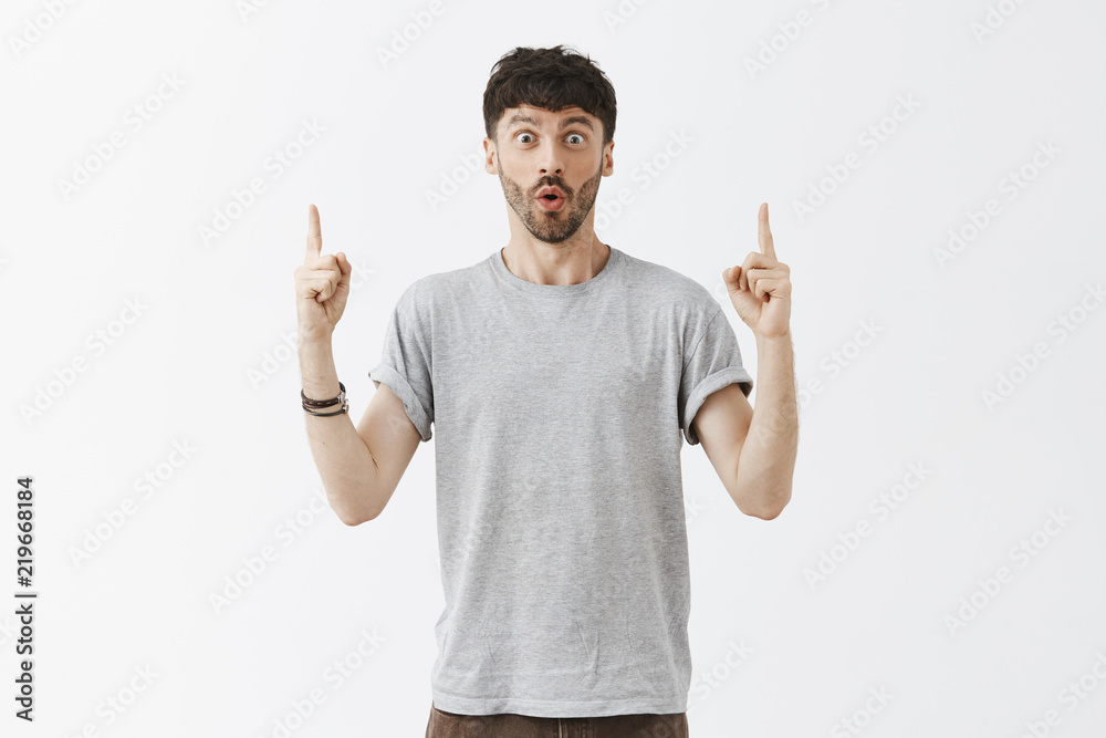 Naklejka premium Guy easy to impress. Portrait of emotive enthusiastic caucasian dark-haired young man with beard and stylish dark hairstyle folding lips with joy, raising index fingers and pointing up with amazement