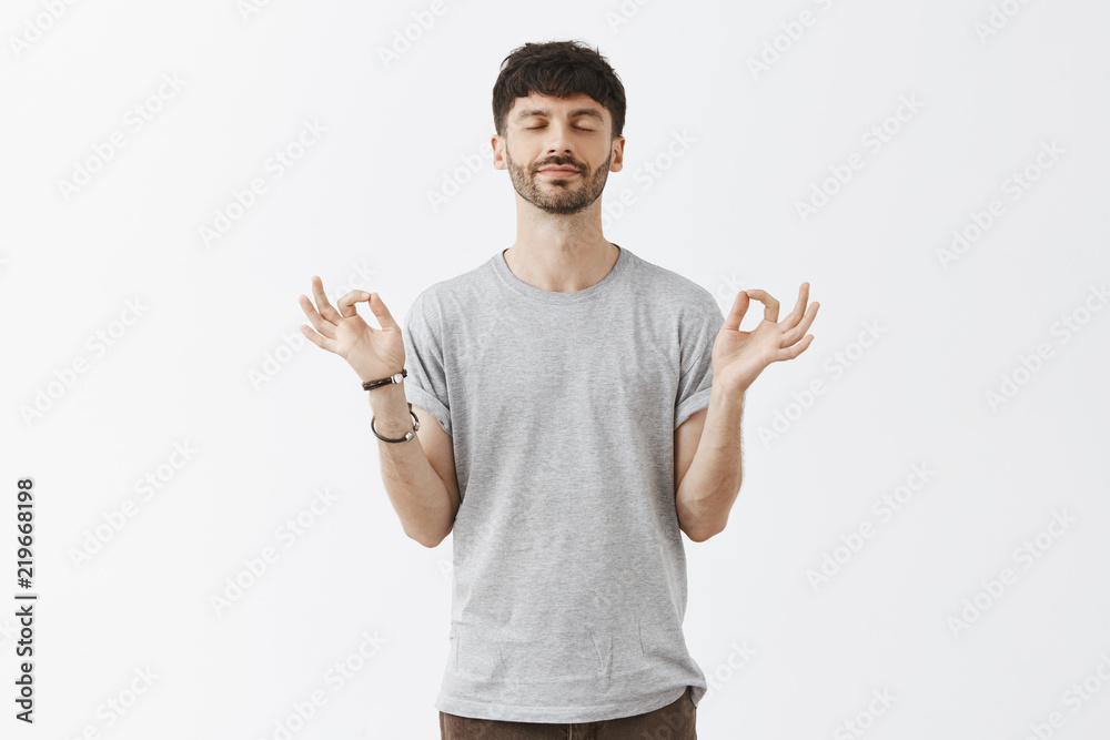 Pacience key to success. Calm and relaxed happy carefree handsome guy with beard in gray casual t-shirt smiling with closed eyes holding hands spread in zen gesture meditating or doing yoga
