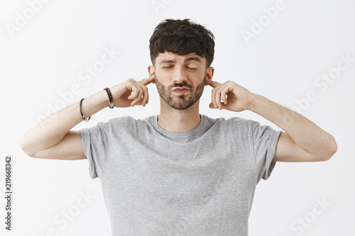 Man prefers not listen to surroundings and enjoy peace inside. Portrait of careless and indifferent calm good-looking male model with beard covering ears smirking and closing eyes over gray wall photo