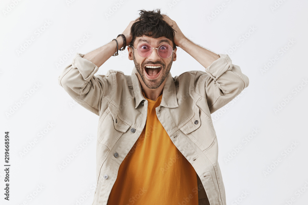 Studio shot of impressed thrilled and excited attractive dark-haired male in stylish sunglasses with beard gasping standing stunned and speechless from happiness and joy holding hands on head Studio shot of impressed thrilled and excited attractive dark-haired male in stylish sunglasses with beard gasping standing stunned and speechless from happiness and joy holding hands on head