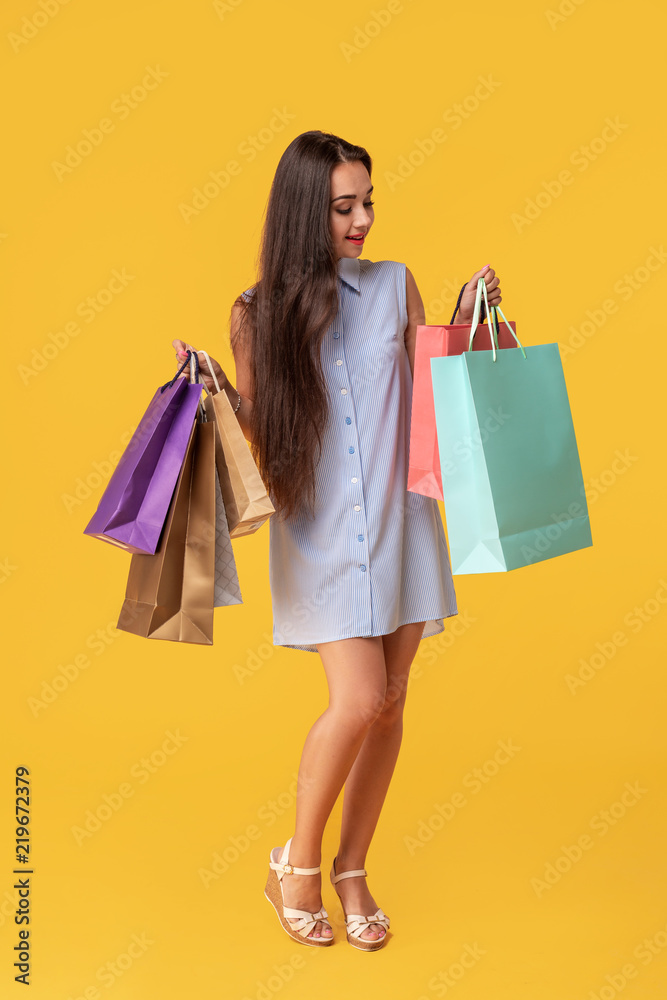Full length photo of young woman in dress looking back on copy space and holding colorful shopping packages over yellow background