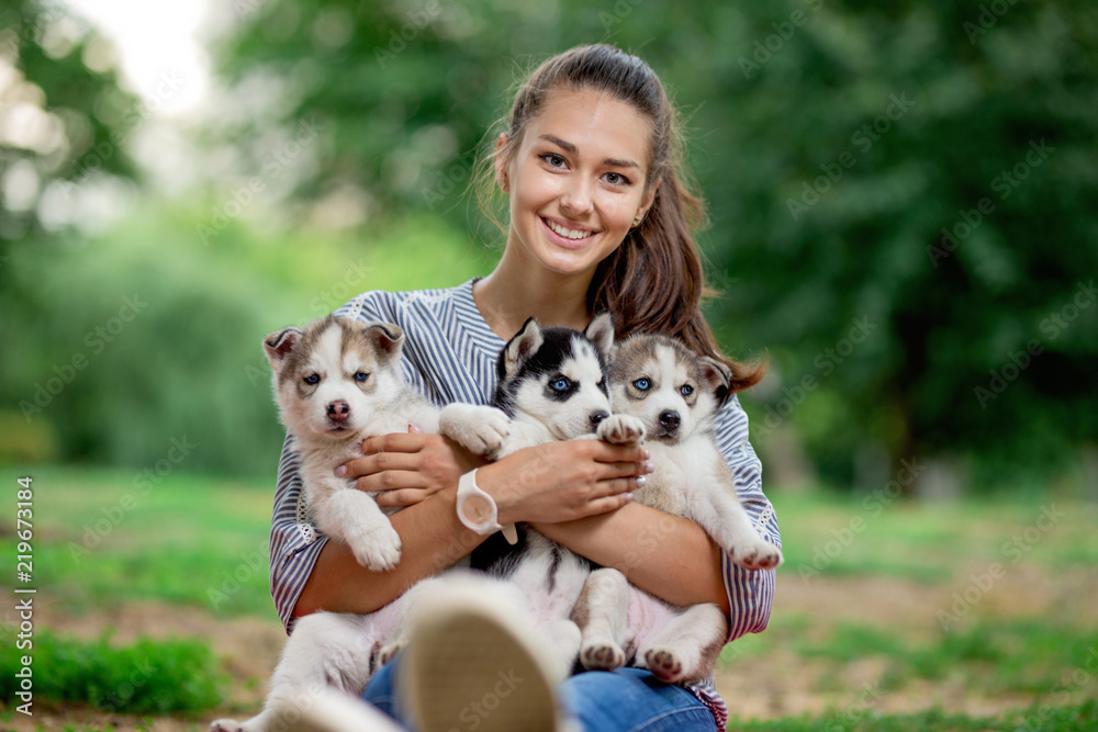 A beautiful smiling woman with a ponytail and wearing a striped shirt is holding three sweet husky puppies on the lawn. Love and care for pets. A beautiful smiling woman with a ponytail and wearing a striped shirt is holding three sweet husky puppies on the lawn. Love and care for pets.