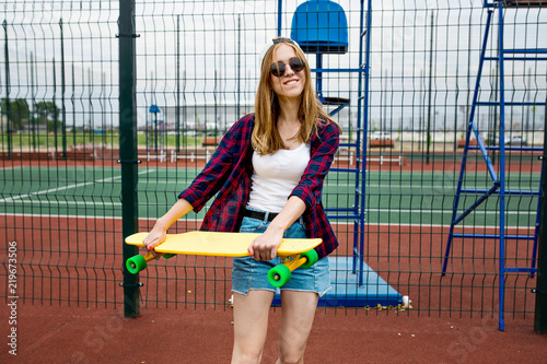 A pretty smiling blond girl wearing checkered shirt  white cap and sunglasses is walking through the sports field with a yellow longboard in her hands. Sport and cool style.