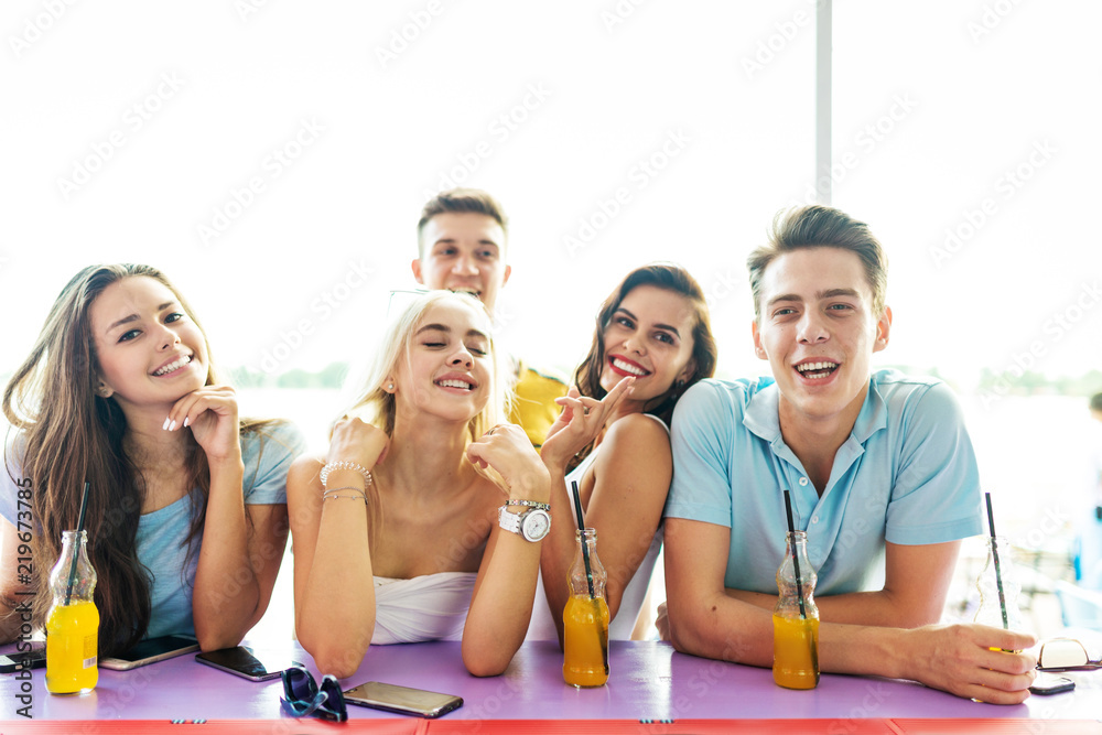 A company of good-looking friends laughing, drinking yellow cocktails and socialising at the bar in the nice summer cafe. River is in the background. Cheers. Entertainment, having good time. 