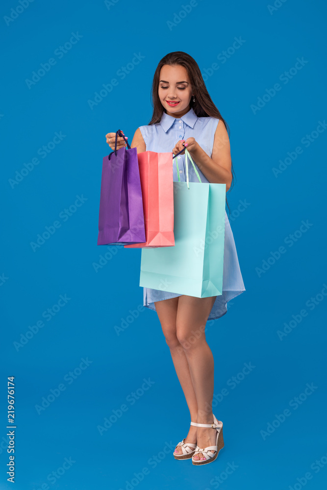 Beautiful woman peeks into a package, in the hands of multi-colored shopping bags on a blue background Beautiful woman peeks into a package, in the hands of multi-colored shopping bags on a blue background
