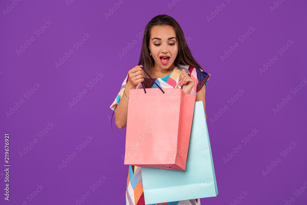 Beautiful woman peeks into a package, in the hands of multi-colored shopping bags on a purple background