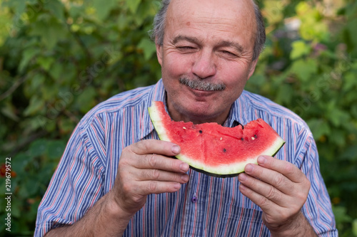 Mature caucasian man with mustache eating juicy water melon with pleasure and smiling.