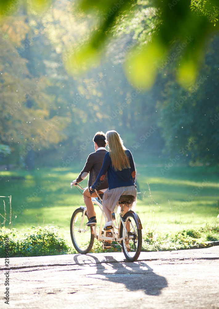 Obraz premium Back view of young couple, man and blond long-haired woman riding tandem double bike over the edge of paved path to green grassy lawn on blurred dense woody forest lit by bright sun background.