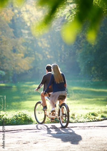 Back view of young couple, man and blond long-haired woman riding tandem double bike over the edge of paved path to green grassy lawn on blurred dense woody forest lit by bright sun background.