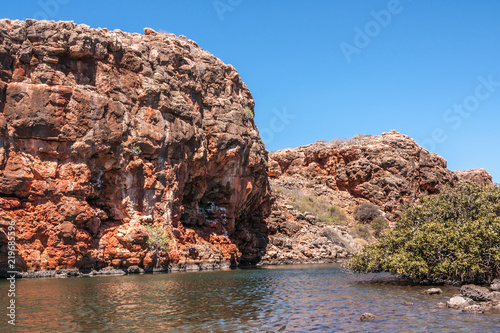 Exmouth, Western Australia - November 27, 2009: Yardie Creek Gorge in Cape Range National Park on North West Cape. Red rock cliffs between gray water and blue sky. Some green vegetation.