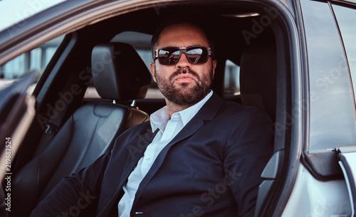 Businessman in sunglasses dressed in a formal suit sitting on the front seats in the luxury car. © Fxquadro
