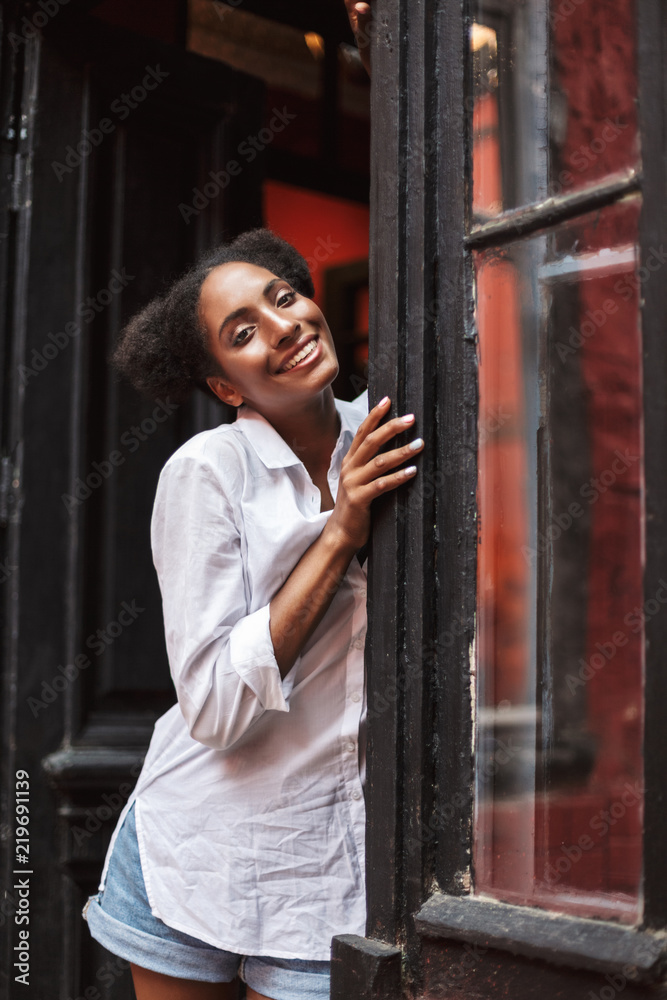 Obraz premium Beautiful smiling african girl with dark curly hair in white shirt joyfully looking in camera while leaning on old black door