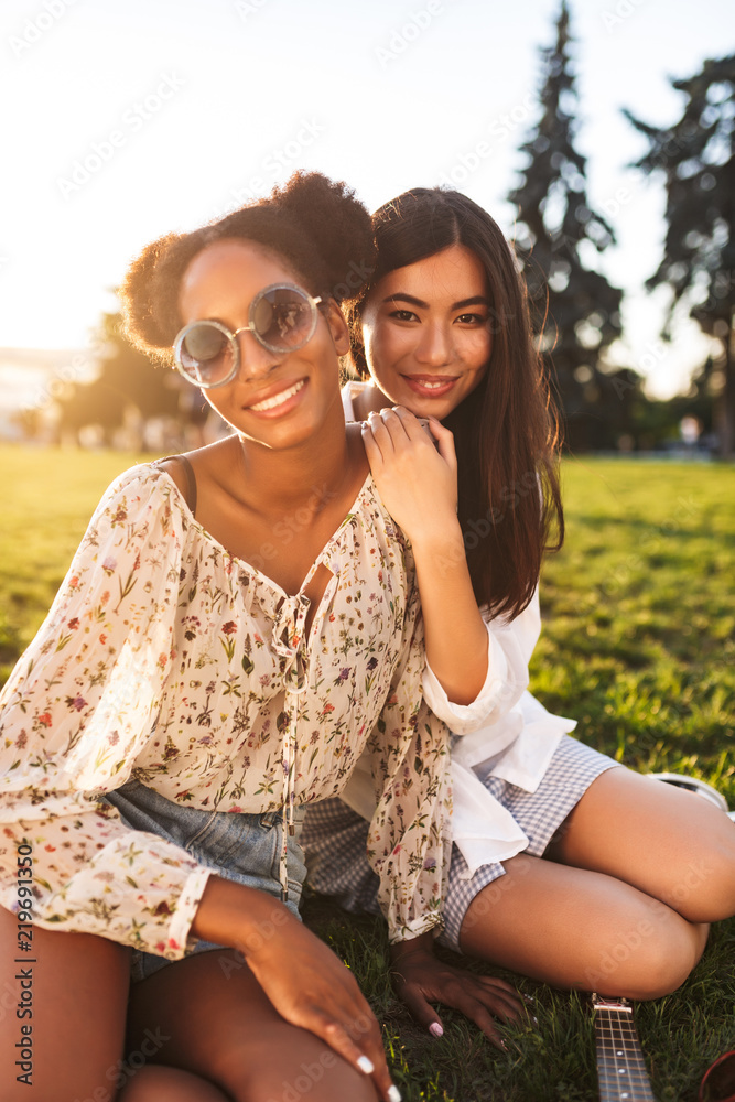 Naklejka premium Two pretty smiling girls sitting on grass joyfully looking in camera while spending time together in city park