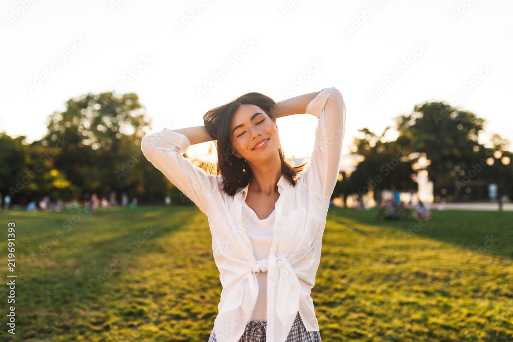 Pretty smiling asian girl in white shirt dreamily closing eyes while happily spending time in park Pretty smiling asian girl in white shirt dreamily closing eyes while happily spending time in park