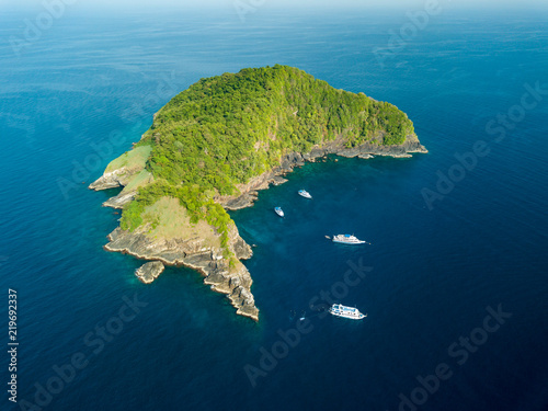 Aerial drone view of boats around a remote, jungle covered tropical island surrounded by fringing coral reef