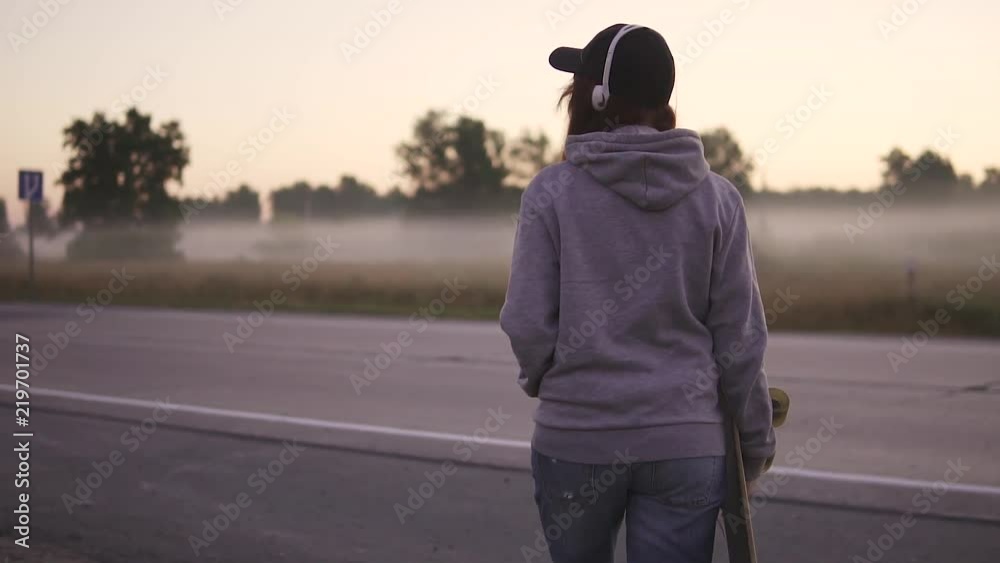 close-up portrait of a hipster girl with a skateboard in hands. The red-haired girl walks along the deserted highway against the background of the morning mist. slow motion. back view close-up portrait of a hipster girl with a skateboard in hands. The red-haired girl walks along the deserted highway against the background of the morning mist. slow motion. back view