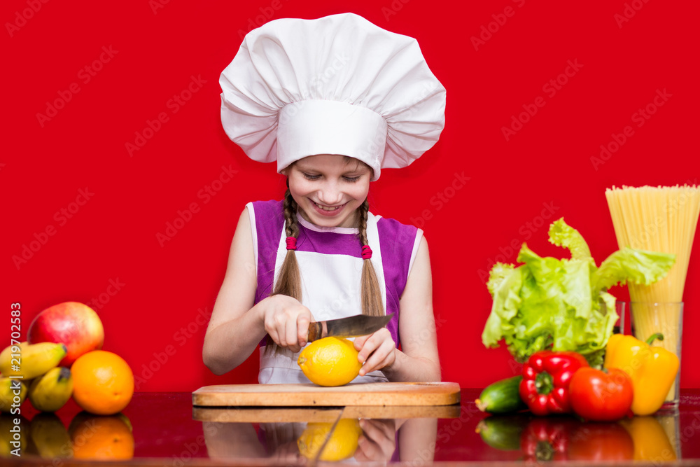 Happy little girl in chef uniform cuts fruit in kitchen. Kid chef. Cooking Process Concept Happy little girl in chef uniform cuts fruit in kitchen. Kid chef. Cooking Process Concept