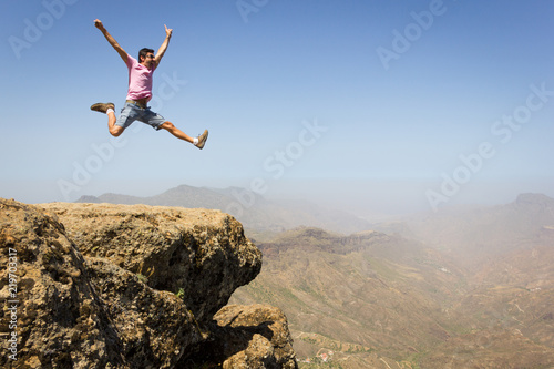 Brave man on pink t shirt jumping high up on rock edge in Roque Nublo natural park, Gran Canaria. Young climber celebrating on mountain peak. Risk, danger, dare, fearless, adventurer, success concepts