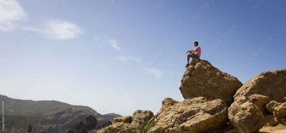 Lonely man sitting on rock top on sunny day in Roque Nublo national park, Gran Canaria. Hiker observing natural landscape panoramic mountain view in Canary Islands, Spain. Explorer, visionary concepts