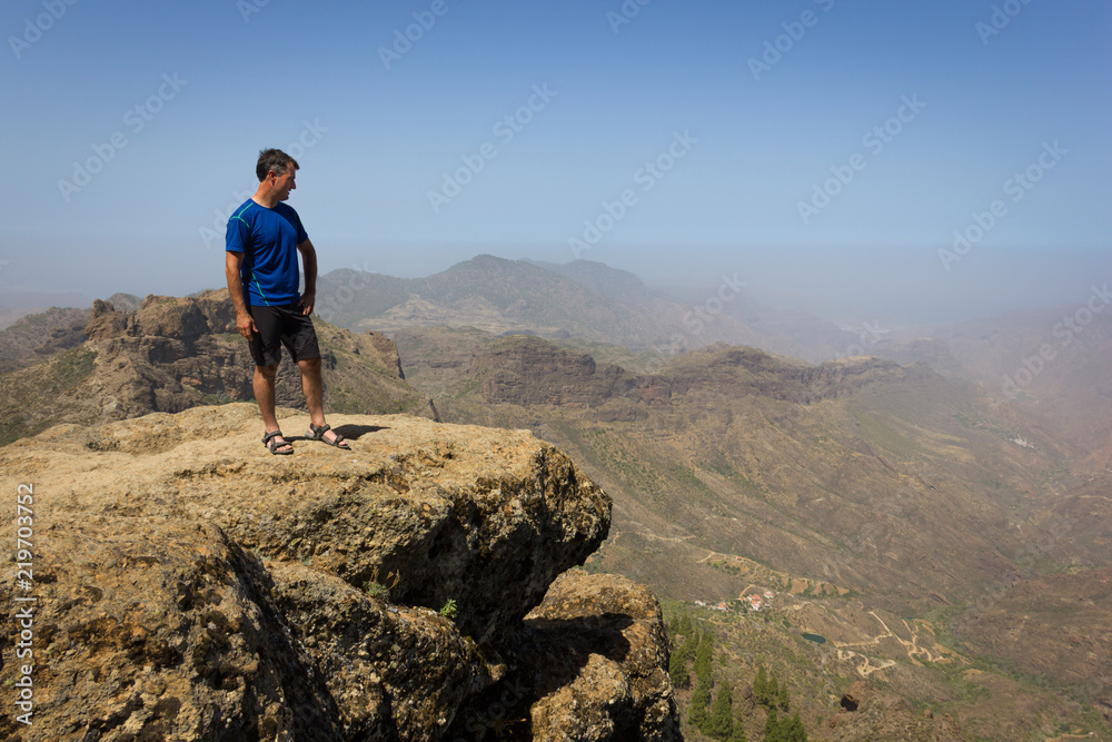 Fototapeta premium Young climber looking down from top of cliff edge in Roque Nublo, Gran Canaria. Adventurer on rocky mountain contemplating panoramic views. Outdoor activity, no fear, explore, freedom concepts