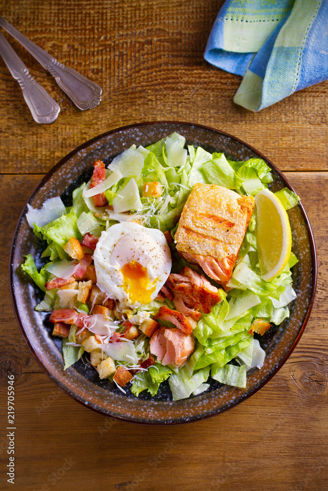 Salmon caesar salad. Crispy pan fried salmon fillet, bacon, poached egg, romaine lettuce and croutons in bowl on wooden table. overhead, vertical