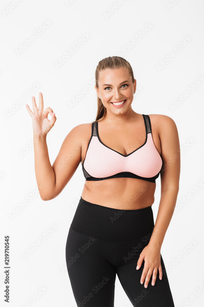 Beautiful plump girl in sporty top and leggings joyfully showing ok gesture while looking in camera over white background. Plus size model Beautiful plump girl in sporty top and leggings joyfully showing ok gesture while looking in camera over white background. Plus size model