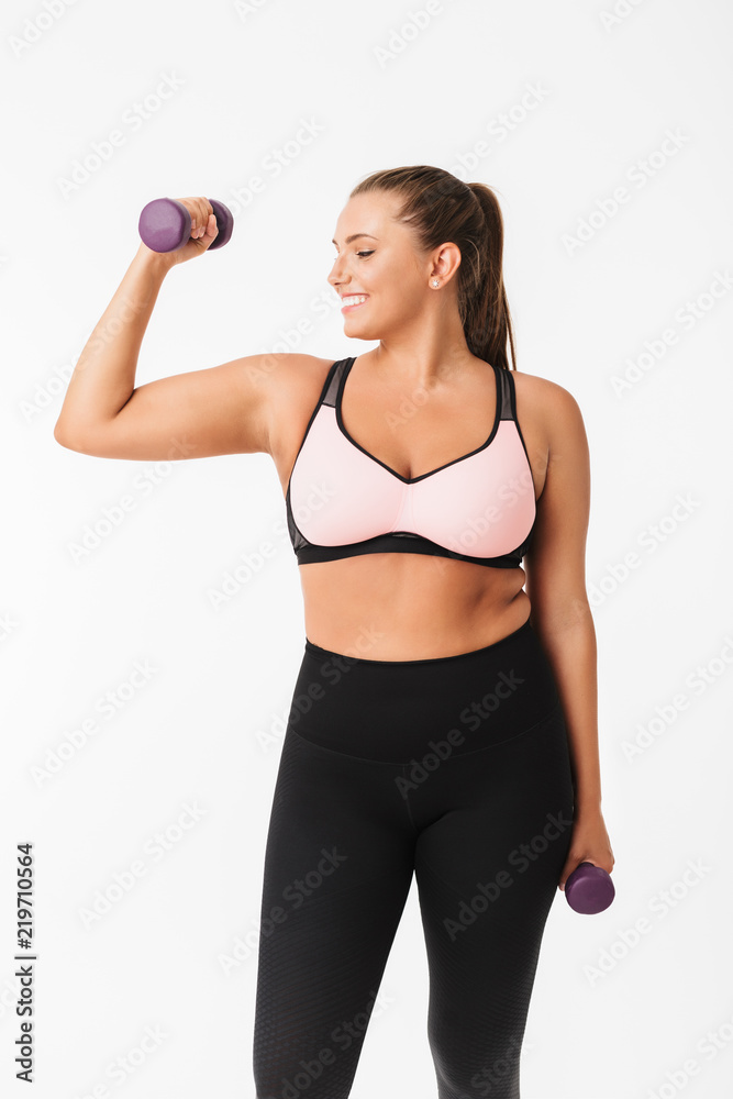 Smiling plump girl in sporty top and leggings holding dumbbells in hands while happily looking aside over white background. Plus size model Smiling plump girl in sporty top and leggings holding dumbbells in hands while happily looking aside over white background. Plus size model