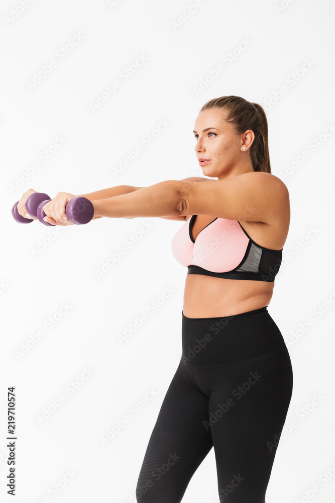 Fototapeta premium Young plump lady in sporty top and leggings doing exercises with dumbbells while dreamily looking aside over white background. Plus size model