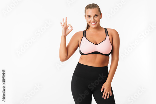 Smiling plump girl in sporty top and leggings happily showing ok gesture while looking in camera over white background. Plus size model