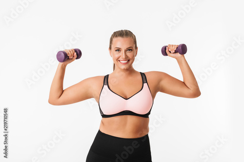 Pretty smiling plump girl in sporty top and leggings holding dumbbells in hands while happily looking in camera over white background. Plus size model