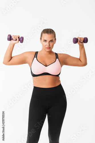 Beautiful plump girl in sporty top and leggings holding dumbbells in hands while thoughtfully looking in camera over white background. Plus size model