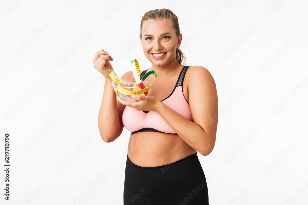 Young woman with excess weight in sporty top pretending that eat measuring tape from bowl while happily looking in camera over white background. Plus size model Young woman with excess weight in sporty top pretending that eat measuring tape from bowl while happily looking in camera over white background. Plus size model
