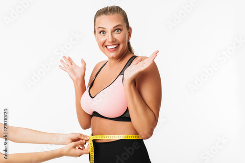 Pretty smiling girl with excess weight in sporty top joyfully looking in camera while measuring waist over white background. Plus size model