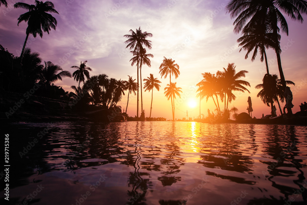 Twilight on a tropical beach with silhouettes of palm trees reflections in water in surreal color.