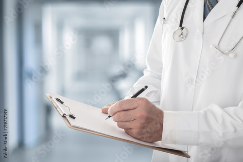Close up of male doctor holding clipboard filling up medical form standing in hospital photo