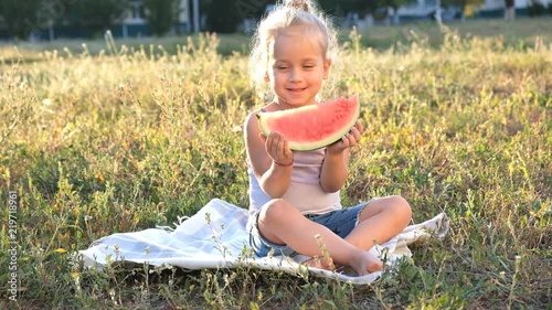 A cute little blonde is sitting in the park on the grass and gaily eating a watermelon. photo