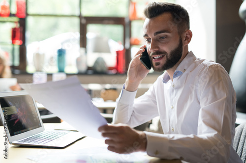 Portrait of handsome bearded businessman wearing white shirt speaking by phone and reading documents while working at table in office or cafe